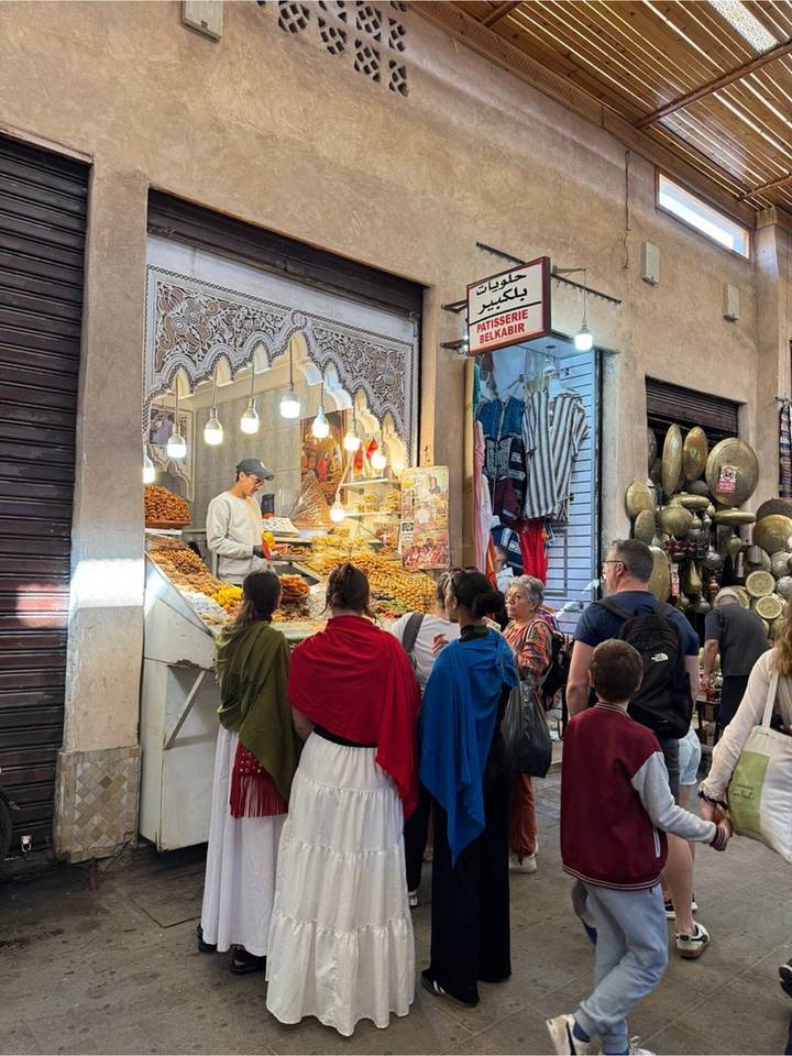 Locals and tourists line up at a patisserie counter filled with Moroccan sweets in a decorated souk passage.