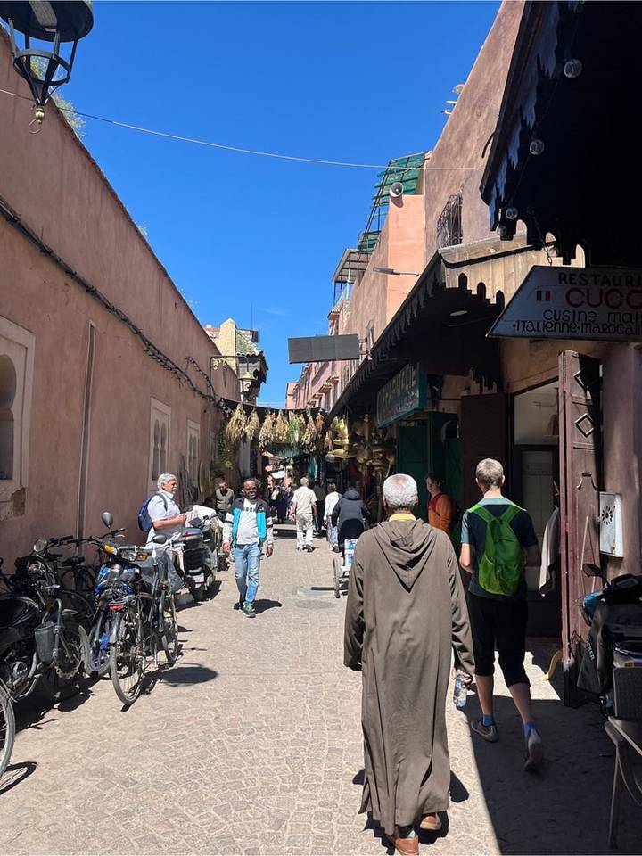 Narrow alleyway with locals and tourists walking between shops and hanging brass lamps in bright sunlight.