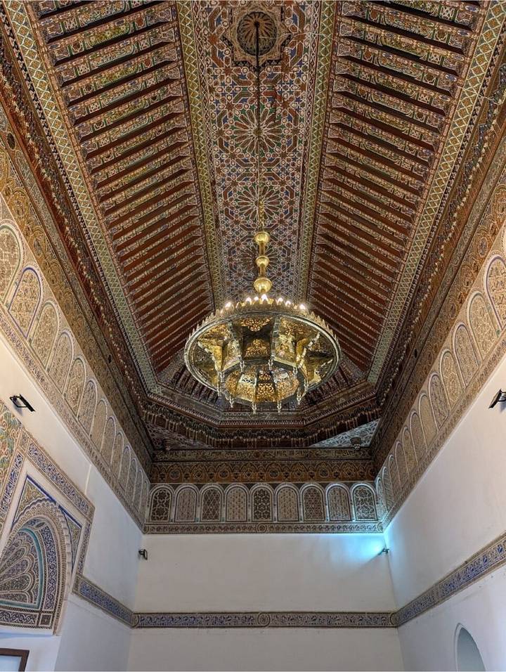 Decorative wooden ceiling with geometric painted patterns and an elaborate gilded chandelier in a Moroccan palace hall.
