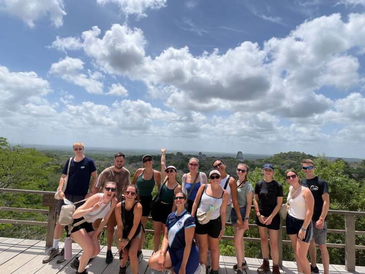 Large tour group poses at a jungle viewpoint overlooking distant Maya temples of Tikal.