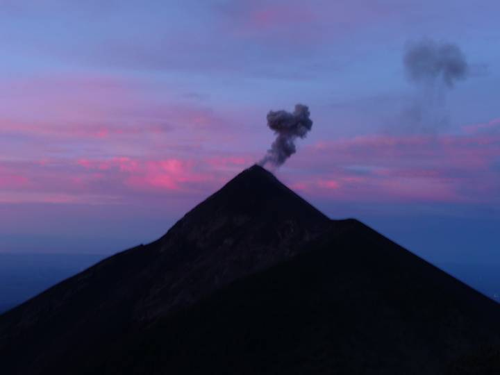 A smoking conical volcano silhouetted against a pink and blue twilight sky.