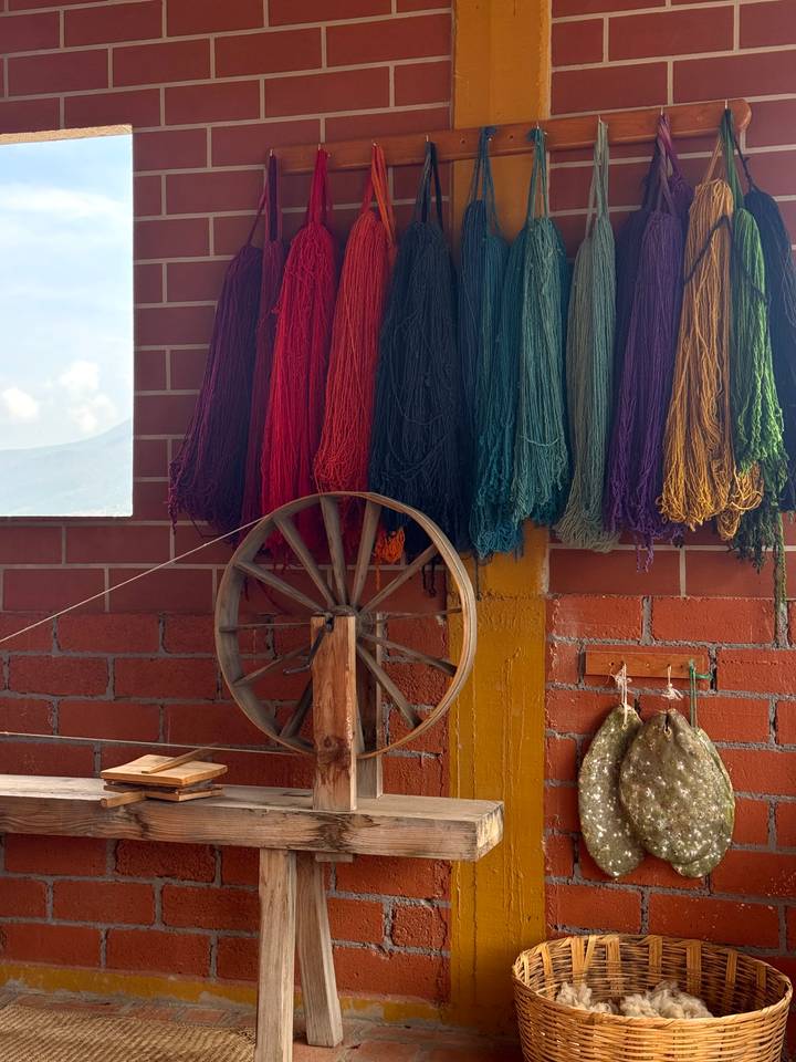 Colorful skeins of yarn hang above a traditional wooden spinning wheel inside a brick-walled workshop.