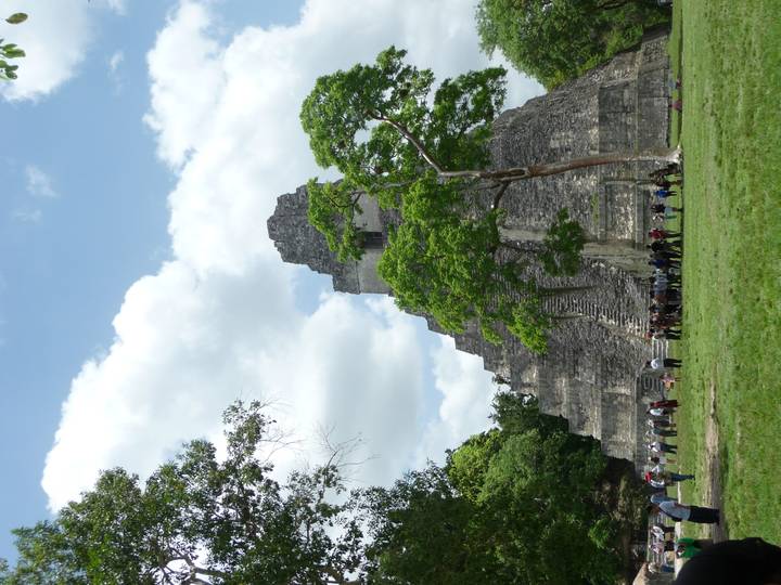 The iconic stepped pyramid of Tikal rises above grassy grounds where tourists gather under a partly cloudy sky.