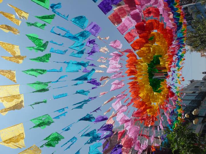 Bright multicolored papel picado banners stretch overhead creating a festive canopy against a clear blue sky.