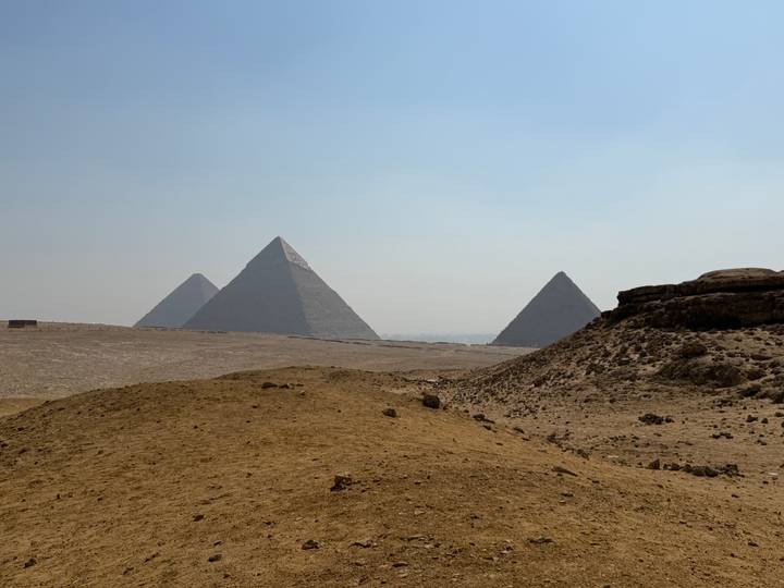 Distant view of the three Pyramids of Giza rising above sandy desert terrain in hazy light