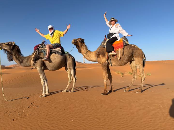 Two travellers pose atop saddled camels in the orange Sahara desert under clear sky.