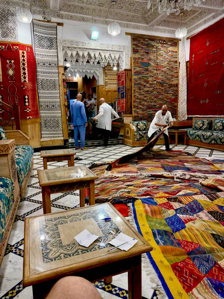 Interior of a carpet shop where men unroll a vividly patterned rug over tiled floor.