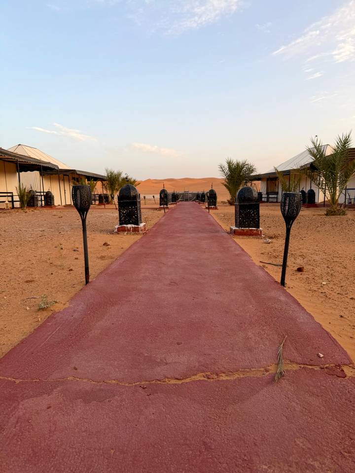 Pathway lined with lantern stands leading to tents in a desert camp with dunes behind.