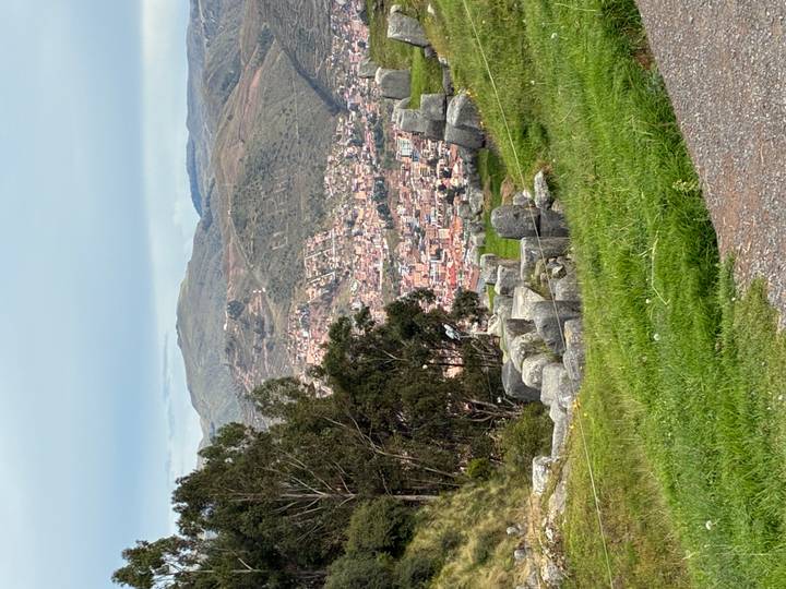 Hillside view over the red-roofed city of Cusco with green mountains and ancient stone ruins in the foreground.