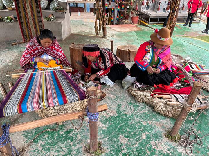 Three Andean women in traditional clothing weave colorful textiles on wooden looms in an open workshop.