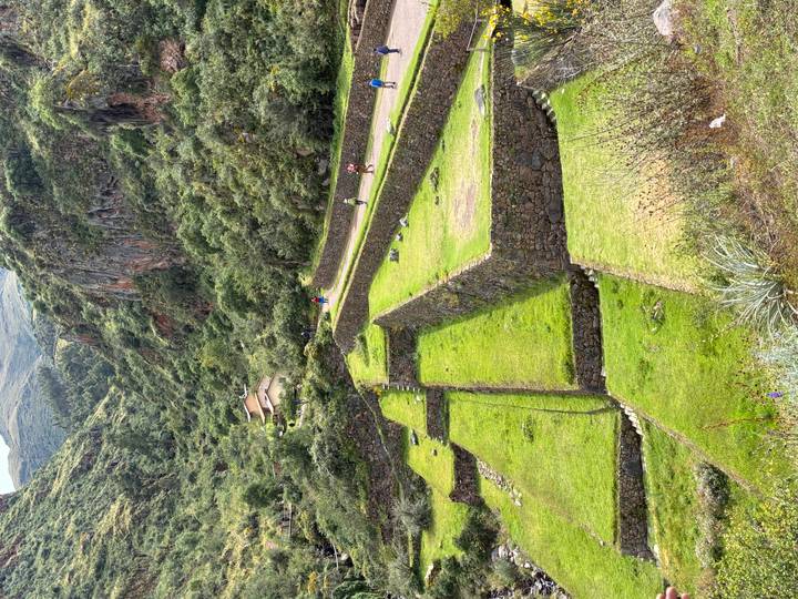 Green terraced fields and stone walls line a winding valley road with hikers in the distance and forested cliffs rising around.