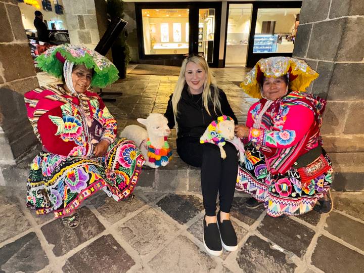 Tourist sitting with two Peruvian women in vibrant traditional attire holding decorated baby alpacas