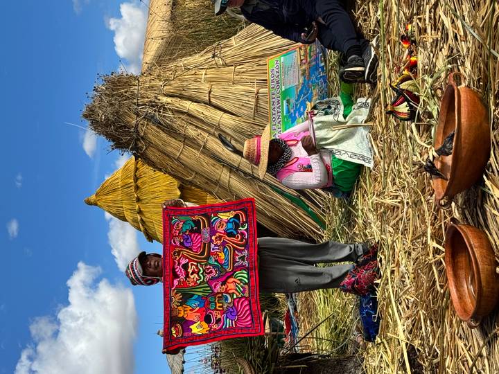 Local man showing colorful textile while woman works beside reed huts on floating Uros Islands