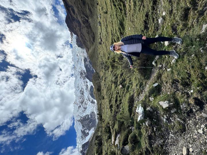 Female trekker with pole on grassy slope beneath snowy Andean peaks and dramatic clouds