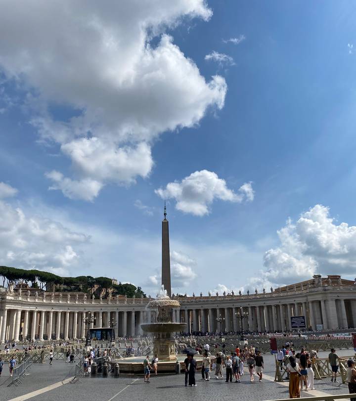 Tall obelisk rises into a bright blue sky framed by fluffy clouds above the colonnades of St. Peter's Square.