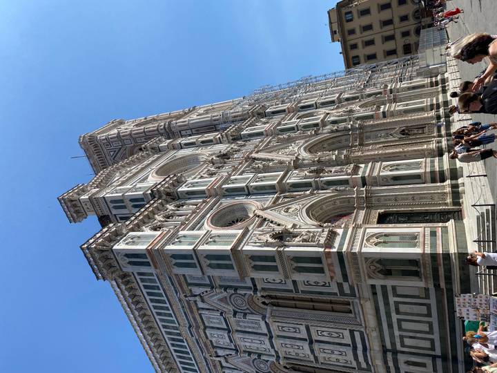 The intricate façade and bell tower of Florence’s Duomo soar against a clear blue sky with visitors below.