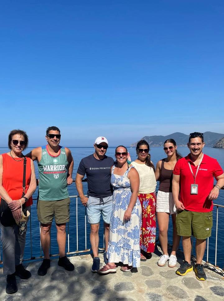 Seven travellers pose on a boat deck with calm blue sea and rugged Ligurian coastline behind.