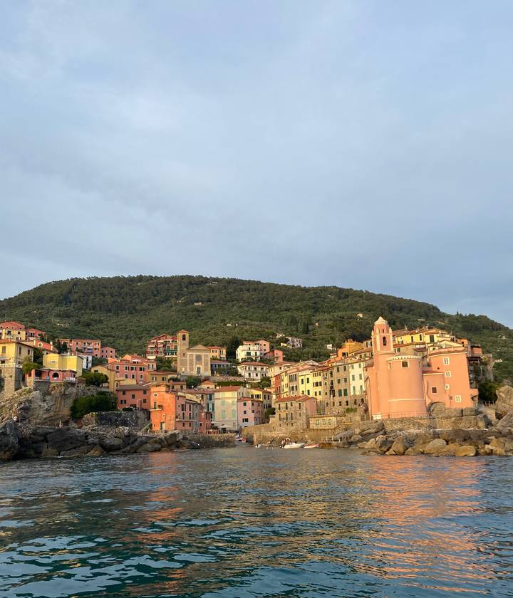Pastel houses cling to a hillside village above the Ligurian Sea under soft evening light.