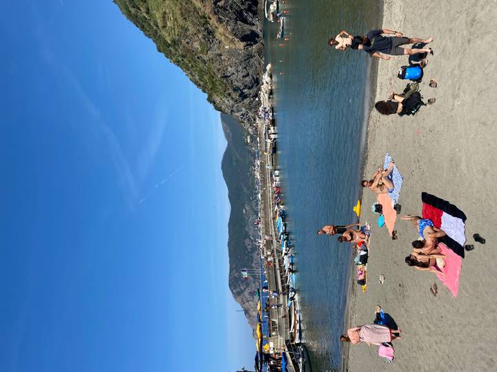 Beachgoers relax on towels beside calm harbour waters bordered by rocky cliffs.