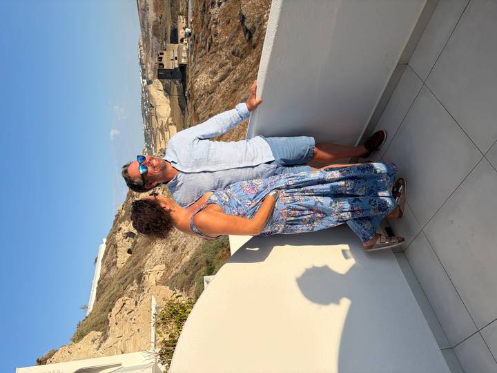 Couple pose on a whitewashed balcony with Santorini’s cliffside village behind in late afternoon light.