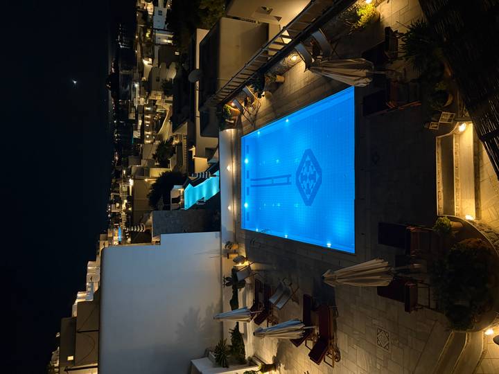 Nighttime view of a glowing turquoise hotel pool and rooftop walkway amid Cycladic buildings.