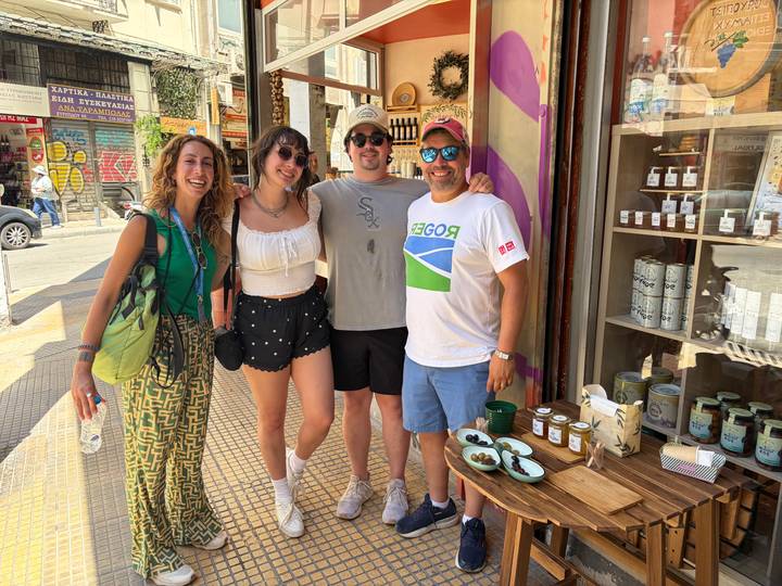 Four friends smile outside a colourful Athens shop selling local goods and olives.