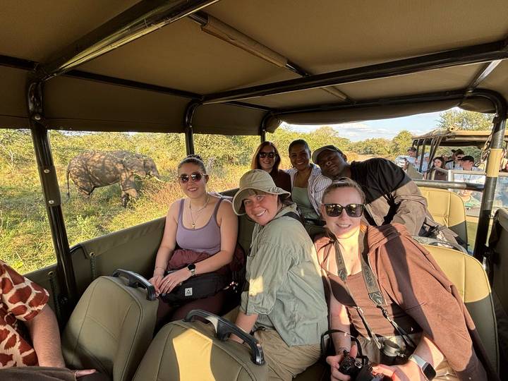 Safari-goers in open jeep smiling while an elephant browses in the bush nearby.