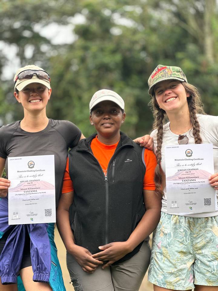 Three climbers proudly display Mount Kilimanjaro summit certificates, smiling for the camera outdoors.
