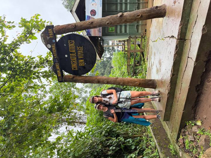Two trekkers with poles pose beneath a wooden Kilimanjaro National Park gate reading 'Congratulations! Bon Voyage'.