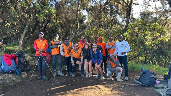 Large team of Kilimanjaro porters and trekkers in orange vests celebrate on a forested mountain trail.