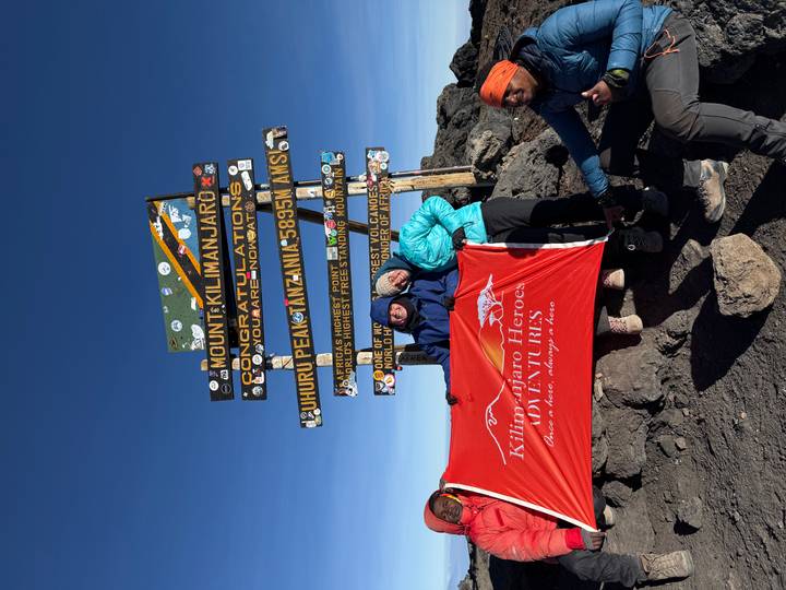 Climbers at Uhuru Peak sign on Mount Kilimanjaro holding a red expedition flag under clear skies.