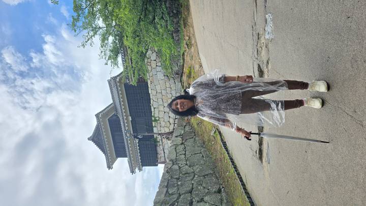 Woman in rain poncho holding umbrella stands on sloping path below a Japanese hilltop castle.