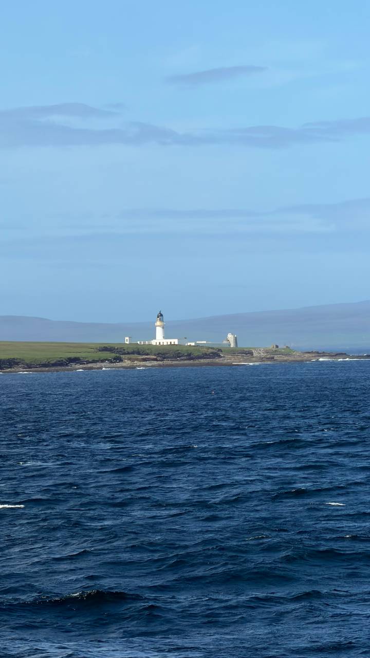 White lighthouse on a grassy headland surrounded by blue sea with hazy hills in the distance