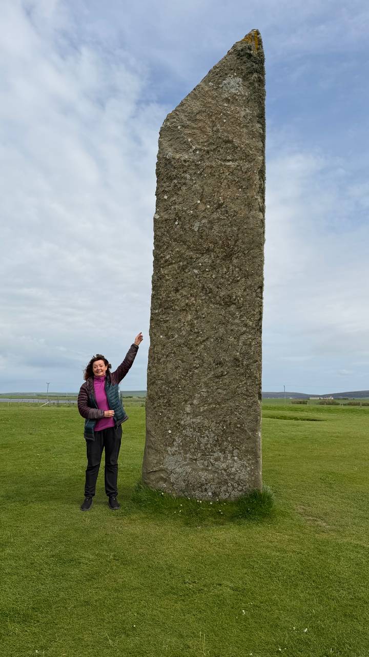 Woman pointing up beside a tall monolithic standing stone on grassy plain