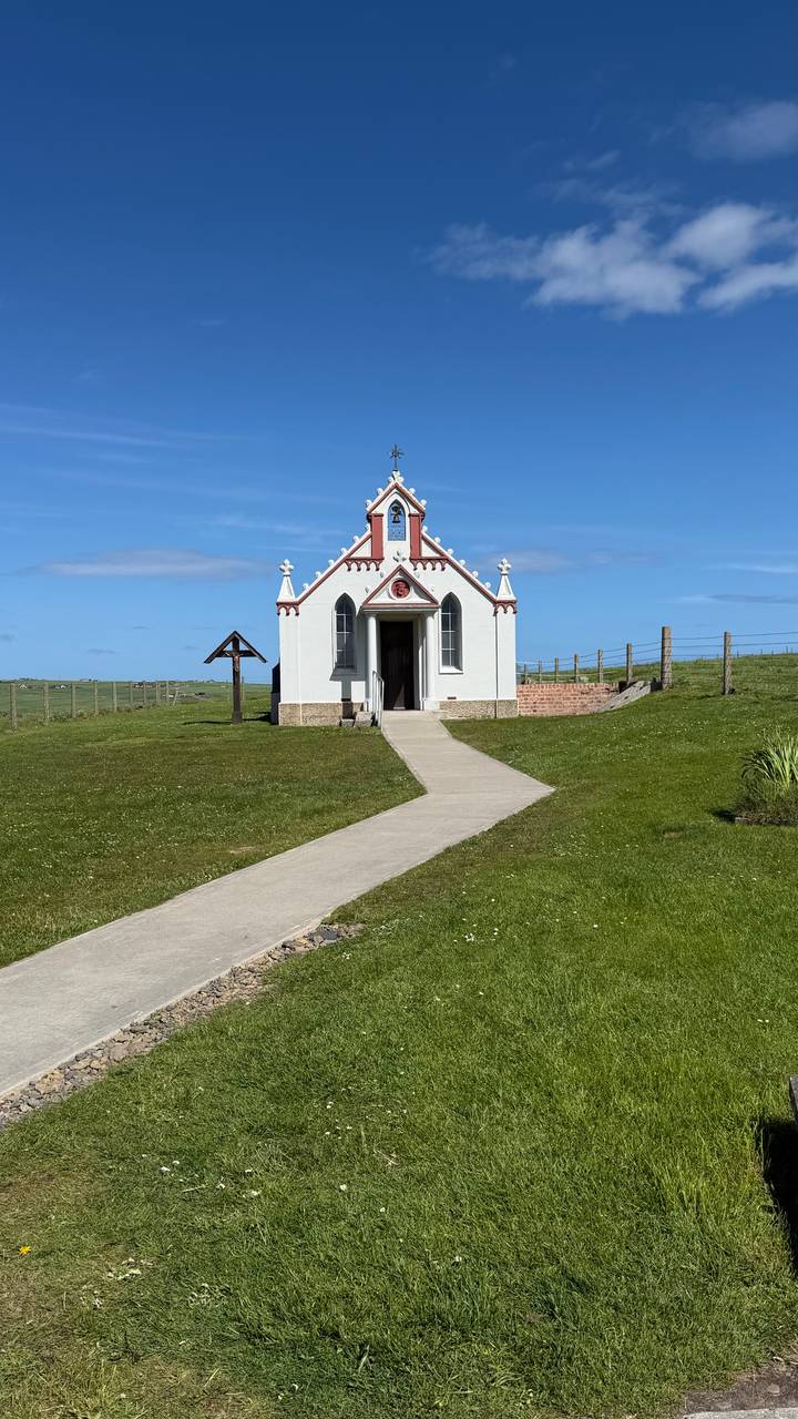 Small white Italian Chapel with red trim standing alone in a grassy field under a bright blue sky