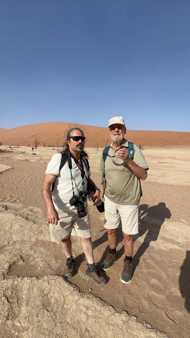 Two men with cameras standing on the cracked white clay pan of Deadvlei against towering red dunes