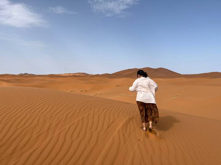 Traveller in white shirt and patterned skirt walks barefoot along rippled desert sands