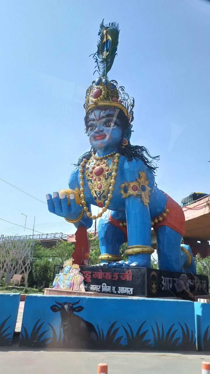 Large blue roadside statue of a jeweled Hindu deity crouching with a fruit in hand against a clear sky.