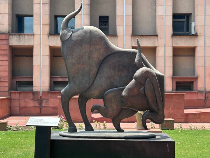 Bronze sculpture of a kneeling bull displayed in a landscaped courtyard outside a modern museum.