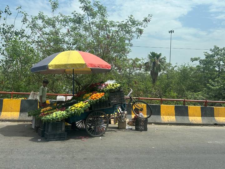 Street vendor sells fresh fruits from a handcart shaded by a multicolored umbrella beside a busy road.