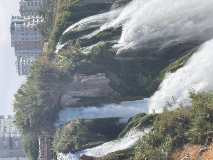 A powerful urban waterfall cascades over a cliff framed by greenery with modern apartment blocks visible on the skyline.