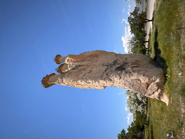 A tall, slender fairy-chimney rock spire rises from a grassy field against a bright blue midday sky.