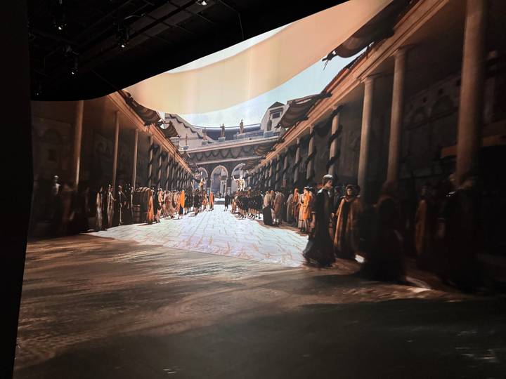 A large immersive screen depicts a bustling ancient marketplace scene inside a dark exhibition space.