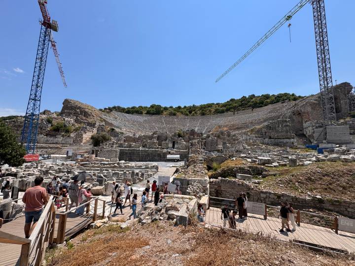 A vast stone amphitheatre under restoration, filled with visitors exploring the ancient seating tiers as cranes loom overhead.