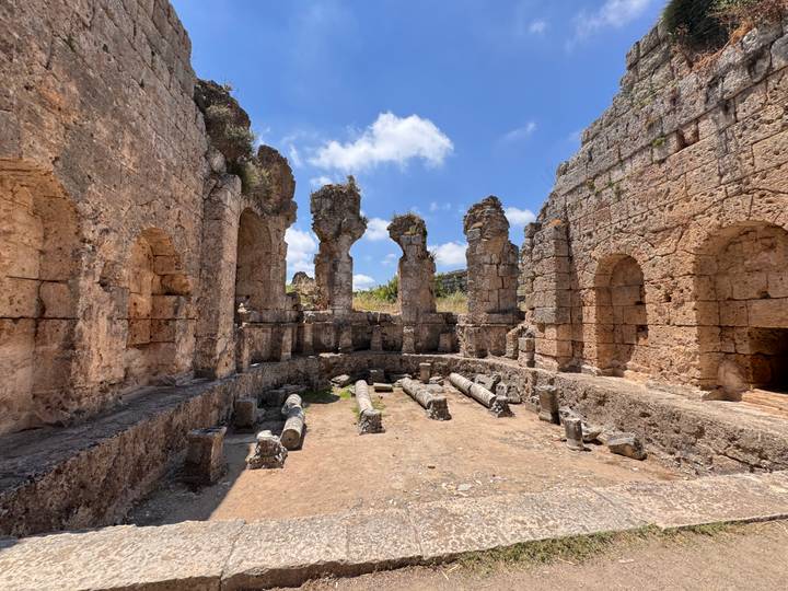 Stone walls and arched niches outline the remains of a small Roman-era theatre or bath complex open to the sky.