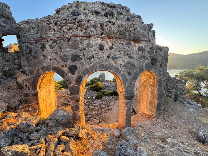 Golden sunset light shines through three weathered stone arches overlooking a tranquil bay and rugged hills.