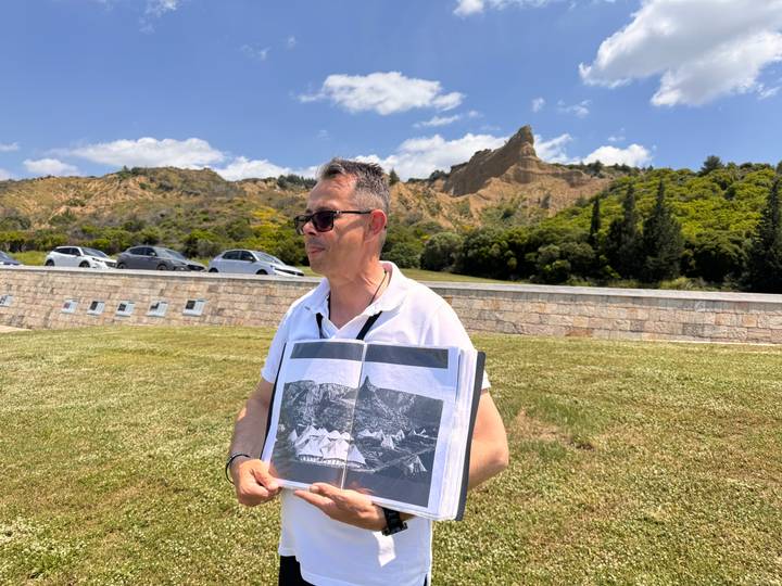 A tour guide stands on a grassy memorial site holding an archival photo while wooded ridges rise behind him.