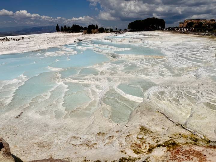 Milky-blue mineral pools and white travertine terraces cascade down a hillside under bright sun.