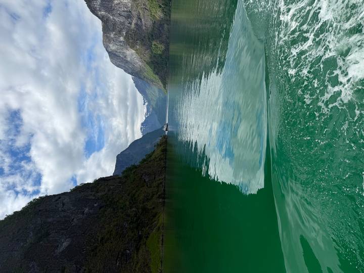 Emerald green fjord waters trail behind a boat between steep cliff walls and snowy peaks