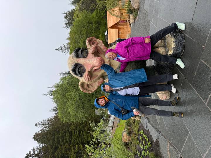 Three travellers in colourful rain jackets pose with a giant whimsical troll statue in a forested park